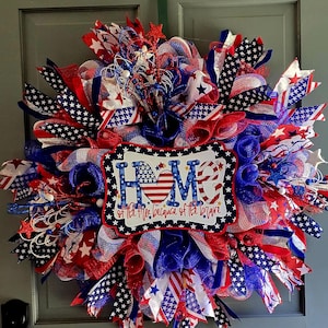 May include: A patriotic wreath in red, white, and blue, featuring stars and stripes. The wreath has a sign that reads "HOME of the free because of the brave". The wreath is made of mesh and ribbons.