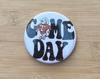 Könnte beinhalten: Ein runder, weißer Button mit den Worten "GAME DAY" in großen, verwitterten schwarzen Buchstaben. Ein Cartoon-Hund mit einem orangefarbenen Hemd mit einem weißen "T" befindet sich in der Mitte. Der Button liegt auf einer Holzoberfläche.