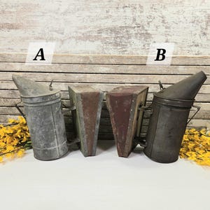 May include: Four antique bee smokers in various styles. Two are cylindrical with angled spouts, and two are rectangular. The smokers are made of metal and have a rustic, weathered appearance. The background is a wooden wall with yellow flowers.