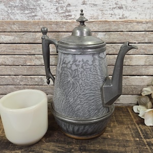 May include: An antique silver-toned coffee pot with a textured gray pattern and a matching cup. The coffee pot has a long spout and a handle. The cup is white and cylindrical. The items are on a wooden surface.