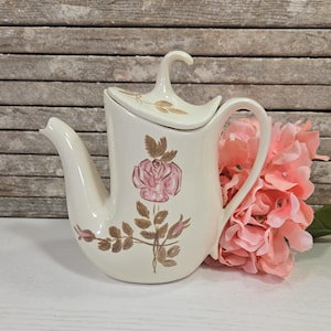Könnte beinhalten: Eine weiße Teekanne aus Keramik mit einem Blumendesign mit rosa Rosen und braunen Blättern. Die Teekanne hat einen gebogenen Ausguss und einen Griff an der Seite.