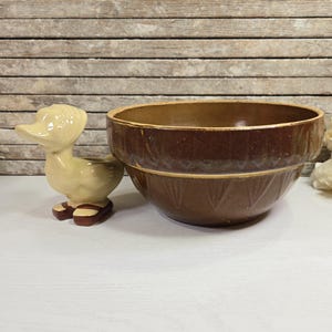 May include: A brown glazed ceramic mixing bowl with a decorative pattern. The bowl is sitting on a white surface next to a small yellow ceramic duck figurine.