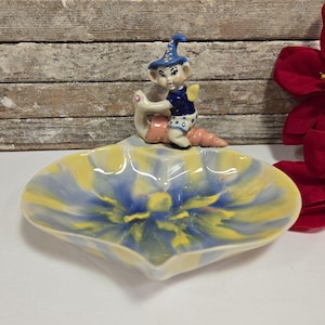 Puede incluir: Un plato de cerámica con forma de flor con un esmalte azul, amarillo y blanco. Una pequeña figura de un hada montando un caracol está posada en el borde del plato.