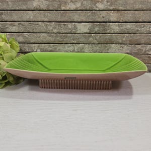 May include: A rectangular, light green ceramic dish with a light brown wooden base. The dish has a smooth, glossy interior and a textured exterior. White lines adorn the corners. The base has a series of vertical wooden slats. Decorative home decor.