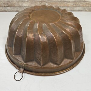 Op de afbeelding: Een vintage koperen cakevorm met een geribbeld ontwerp en een ophanglus. De vorm is in goede staat met een patina.
