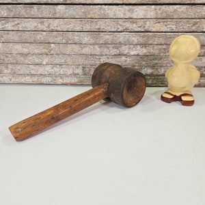 May include: A wooden mallet with a brown wooden handle and a dark brown, round head. The mallet is sitting on a white surface.