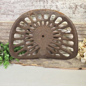 Könnte beinhalten: Ein Vintage-Traktorsitz aus Gusseisen mit halbkreisförmiger Form und radialem Speichendesign. Der Sitz hat eine verwitterte, rostbraune Oberfläche und ein zentrales quadratisches Loch. Der Hintergrund zeigt Holzbretter und Grün.