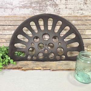 Könnte beinhalten: Ein dunkelbraunes, halbkreisförmiges Metallobjekt mit ovalen und kreisförmigen Aussparungen. Das Objekt ist wahrscheinlich ein Vintage- oder antikes Sitz- oder Dekorationsteil. Ein klares, blau getöntes Glasgefäß ist im Hintergrund zu sehen.