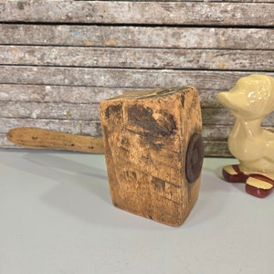May include: A wooden mallet with a worn handle and a metal ring on the head. The mallet is resting on a light gray surface.