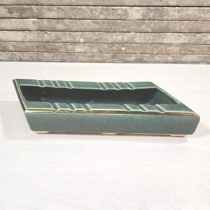 Könnte beinhalten: Ein rechteckiger Aschenbecher in gedämpftem Teal mit goldfarbenem Rand. Der Aschenbecher hat eine vertiefte Mitte und mehrere Rillen zum Ablegen von Zigaretten. Der Sockel ist hellbeige.