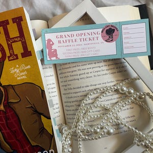Puede incluir: Una imagen de naturaleza muerta con un libro titulado "Lucky River Ranch Novel" y un boleto de rifa para Bellamy Brooks. El boleto es azul claro con acentos rosas y enumera los premios. También hay un collar de perlas y un libro abierto.