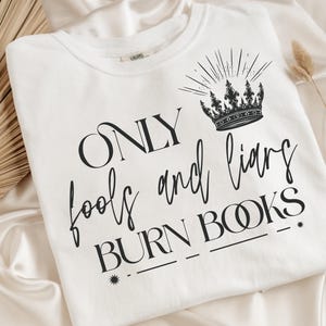 May include: White t-shirt featuring the text "ONLY fools and liars BURN BOOKS" in black script, along with a crown graphic. The shirt is displayed on a satin surface.