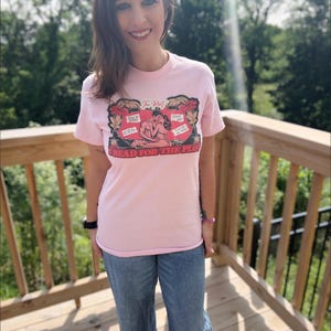 May include: A light pink short-sleeve t-shirt featuring a graphic illustration of a couple embracing. The design includes the text "The Plot" and "READ FOR THE PLOT" along with speech bubbles containing the phrases "You're mine," "Good girl," "It'll fit," and "It's me, baby."