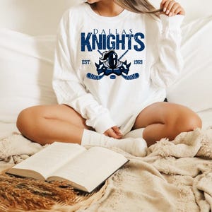 Puede incluir: Sudadera blanca con un logotipo de Dallas Knights azul y blanco. El logotipo presenta un casco de caballero con palos de hockey cruzados y el texto "Dallas Knights" y "Est. 1969".
