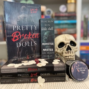 May include: A stack of books titled "Pretty Broken Dolls," "Pretty Stolen Dolls," "Pretty Lost Dolls," and "Pretty New Dolls." A bookmark with a skull and the text "Monster and Master are ready to play." A skull and a candle are also present.