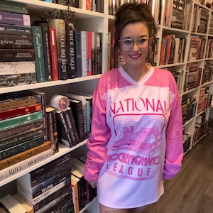May include: A pink and white long-sleeved shirt with the text "NATIONAL HOCKEY ROMANCE LEAGUE" in a retro design. The shirt features a V-neck and is worn in front of a bookshelf filled with books.