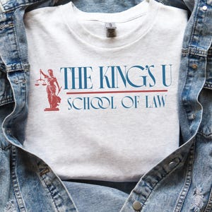 Puede incluir: Camiseta gris claro con el texto "THE KING'S U SCHOOL OF LAW" en azul y rojo. Una ilustraci&oacute;n roja de una mujer con una balanza est&aacute; a la izquierda. La camiseta se muestra sobre una chaqueta vaquera.