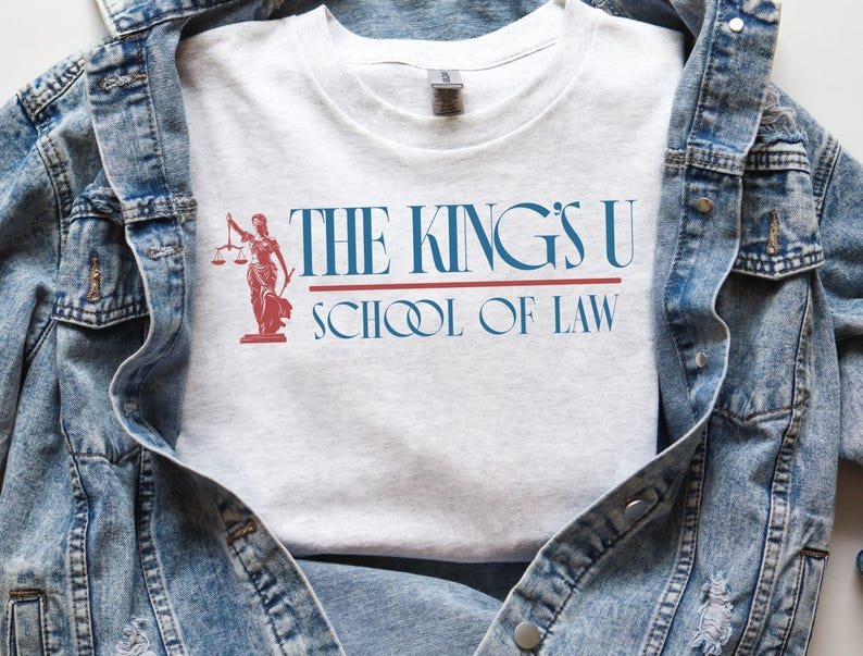 Puede incluir: Camiseta gris claro con el texto "THE KING'S U SCHOOL OF LAW" en azul y rojo. Una ilustraci&oacute;n roja de una mujer con una balanza est&aacute; a la izquierda. La camiseta se muestra sobre una chaqueta vaquera.