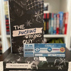 May include: Stacked books with the titles "The Pucking Wrong Guy", "The Pucking Wrong Date", and "The Pucking Wrong Number". A hockey ticket for Knights vs Cobras is on top. The books are black with white text.