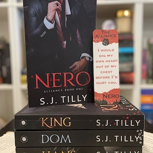 Op de afbeelding: Een stapel boeken met de titels "King", "Dom" en "Hans" van S.J. Tilly. Het boek "Nero" ligt bovenop, met een bladwijzer met een citaat en de titel "Nero".