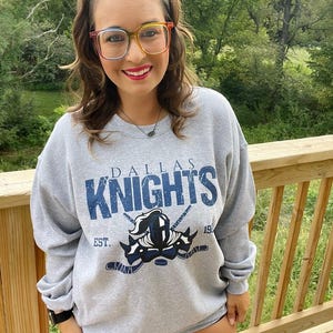 Puede incluir: Sudadera gris con un logotipo de Dallas Knights azul y blanco. El logotipo presenta un casco de caballero con palos de hockey cruzados y el texto "Dallas Knights" y "Est. 19."
