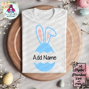 Könnte beinhalten: Weißes T-Shirt mit blauem Osterei-Design und rosa Hasenohren. Der Text "Add Name" befindet sich unter dem Ei. Das Shirt liegt auf einem Holztablett mit Ostereiern und floralen Akzenten. Digitaler Download.