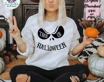 May include: A cream-colored sweatshirt with a black Halloween-themed graphic that reads "This is Halloween." The design features a stylized face with the text incorporated into the design. The image also includes a small orange pumpkin.