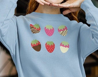 Puede incluir: Una sudadera de color azul claro con un gráfico de seis fresas, cada una con una cobertura de chocolate diferente. Las fresas están dispuestas en dos filas de tres.