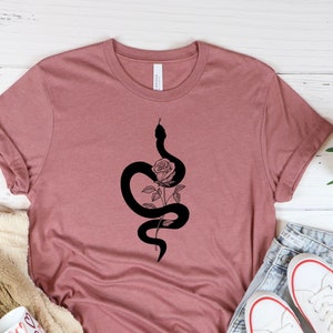 Peut inclure: Un t-shirt rose poussiéreux avec un graphique noir d'un serpent enroulé autour d'une rose. Le t-shirt est posé sur une surface blanche avec une couverture et une paire de baskets blanches.