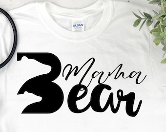 Puede incluir: Camiseta blanca con texto negro que dice "Mama Bear" y una huella de pata de oso. La camiseta está sobre una superficie de madera con pantalones vaqueros azules y zapatillas rojas.