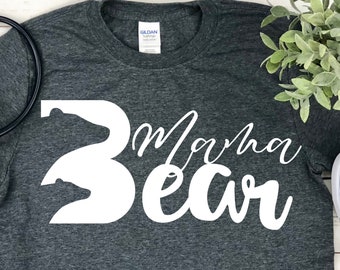 Puede incluir: Una camiseta gris oscuro con una silueta de oso blanca y el texto "Mama Bear" en letras blancas cursivas. La camiseta se combina con jeans azules y zapatillas rojas.