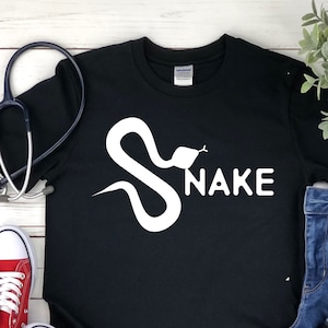 May include: Black t-shirt featuring a white snake graphic and the word "SNAKE" printed on it. The shirt is laid out on a white wooden surface with a stethoscope, red trainers, blue jeans, and a black tumbler.