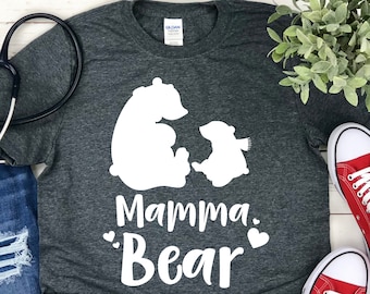 Puede incluir: Una camiseta gris oscuro con un gráfico blanco de dos osos. El oso más grande está sentado con sus brazos alrededor de un oso más pequeño. El texto "Mamma Bear" está debajo de los osos con dos corazones blancos.