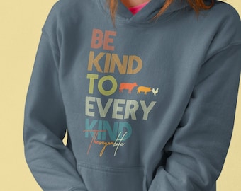 May include: A dark blue hooded sweatshirt with the text "Be Kind To Every Kind" in a rainbow color scheme. The text is in a vintage style font. The text is centered on the sweatshirt. The sweatshirt is being worn by a person with red hair.