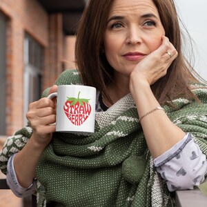 May include: A white ceramic mug with a red strawberry design. The word "Strawberry" is written in a stylized font inside the strawberry shape.