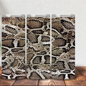 May include: Three cylindrical tumblers featuring a continuous brown, black, and white snakeskin pattern. Each tumbler has a smooth, straight-walled design with a metallic rim at the top, showcasing a detailed reptilian scale texture throughout.
