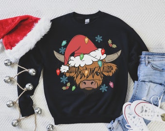 May include: A black crewneck sweatshirt featuring a graphic of a brown Highland cow wearing a red Santa hat. The cow is decorated with colorful Christmas light bulbs and surrounded by light blue snowflakes and small sprigs of holly berries.