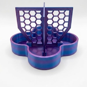 May include: A clover-shaped storage tray featuring a central vertical divider with a honeycomb lattice pattern. The object is constructed from a dual-tone filament, displaying alternating horizontal bands of vibrant purple and blue throughout the structure.