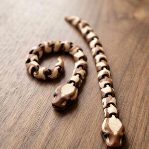 May include: Two articulated wooden snake figures featuring a segmented design with alternating light and dark brown wood grain patterns. Each snake has a carved head with small dark eyes and a flexible body composed of interlocking wooden pieces.