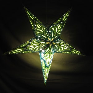 May include: A large, illuminated star-shaped paper lantern. The lantern features a detailed, symmetrical design with green and white patterns. The star is suspended against a dark background, creating a striking contrast. The lantern is lit from within, casting a warm glow.
