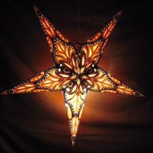 May include: A five-pointed star-shaped paper lantern with intricate black and orange floral designs. The lantern is illuminated from within, casting a warm glow.