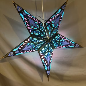 May include: A five-pointed star-shaped hanging lamp featuring intricate cut-out patterns. The structure is black with gold-toned edges, revealing a vibrant inner glow of bright cyan and soft pink light through the decorative floral-inspired cut-outs.