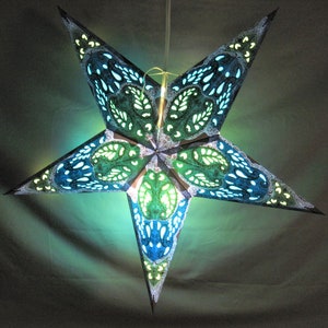 May include: A teal and green paper star lantern with a white lace design. The star is illuminated from within, casting a warm glow.