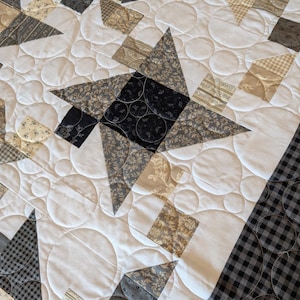 May include: Close-up of a handmade quilt with star and geometric patterns. The quilt features fabrics in shades of grey, black, and beige, with white accents and circular quilting. The border has a black and white checkered pattern.