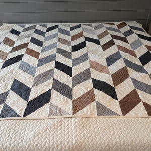 May include: A handmade quilt with a chevron pattern in shades of black, brown, grey, and cream. The quilt is placed on a bed and features intricate stitching details. The neutral colour palette makes it suitable for various bedroom decor styles.