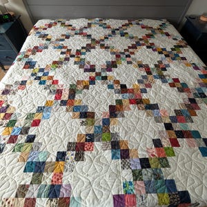 May include: A quilt featuring a white background with a repeating lattice pattern composed of small, multi-coloured fabric squares in various prints and solid colours. The entire surface is finished with a continuous, swirling machine-stitched design.