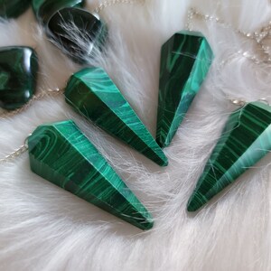 May include: Four green malachite crystal pendulums with silver chains on a white fluffy surface.