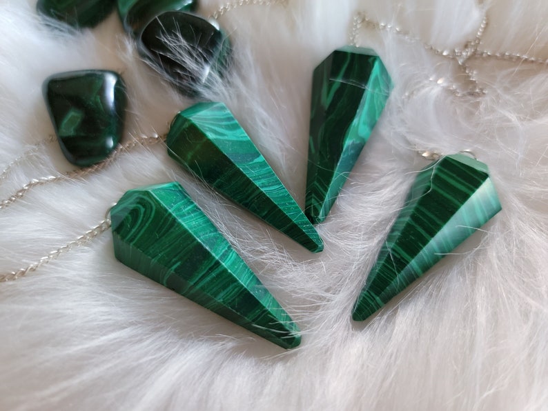 May include: Four green malachite crystal pendulums with silver chains on a white fluffy surface.