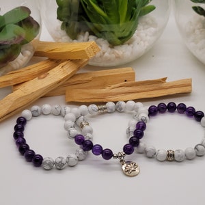 May include: Three beaded bracelets with white and purple stones. One bracelet has a silver charm. The bracelets are displayed with Palo Santo sticks and decorative glass terrariums with plants.