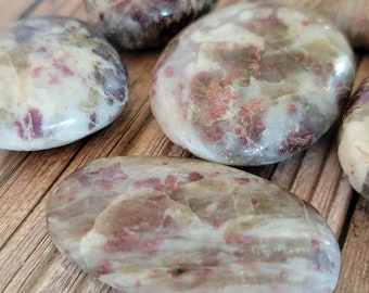 May include: A collection of five smooth, oval-shaped stones with a white base and red and brown flecks. The stones are arranged on a wooden surface.